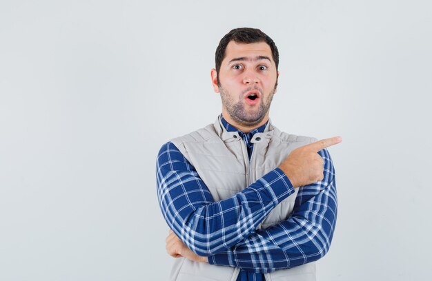 Young man pointing to left side in shirt,sleeveless jacket and looking surprised. front view.