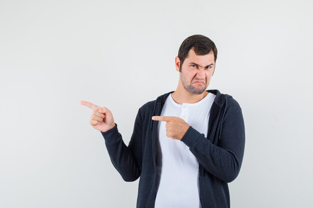 Young man pointing left with index fingers in white t-shirt and zip-front black hoodie and looking displeased , front view.