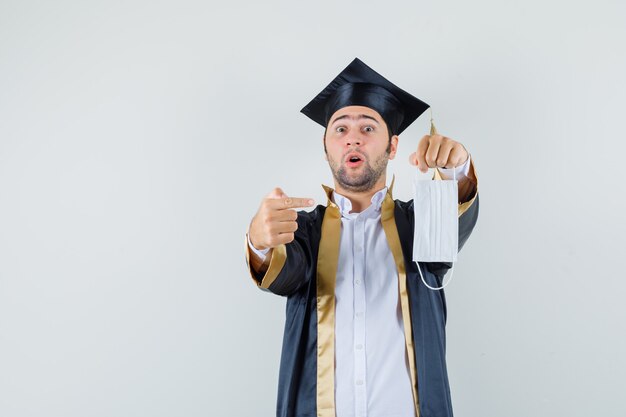 Young man pointing at medical mask in graduate uniform and looking surprised , front view.