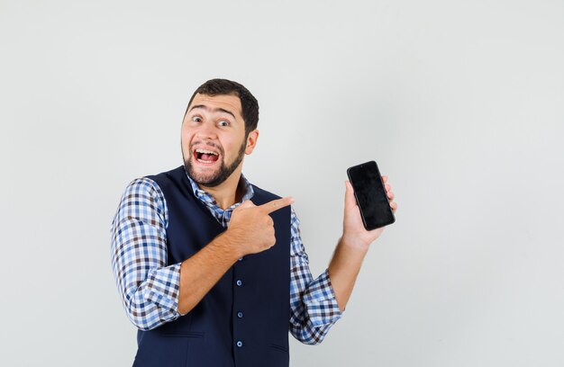 Young man pointing at mobile phone in shirt, vest and looking happy.