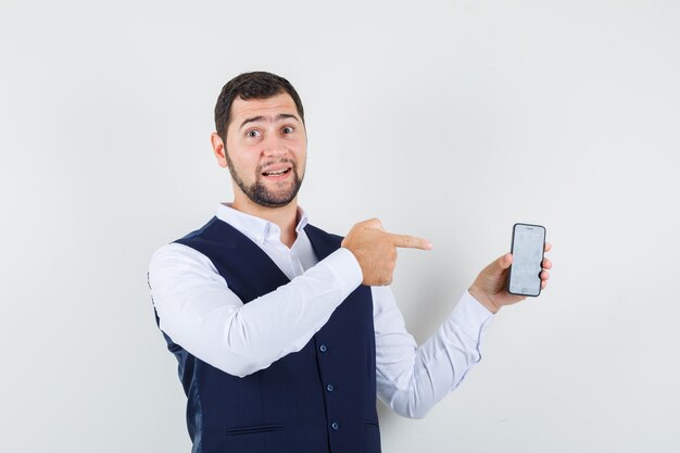 Young man pointing at mobile phone in shirt, vest and looking pleased