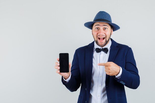 Young man pointing at mobile phone in suit, hat and looking happy. front view.