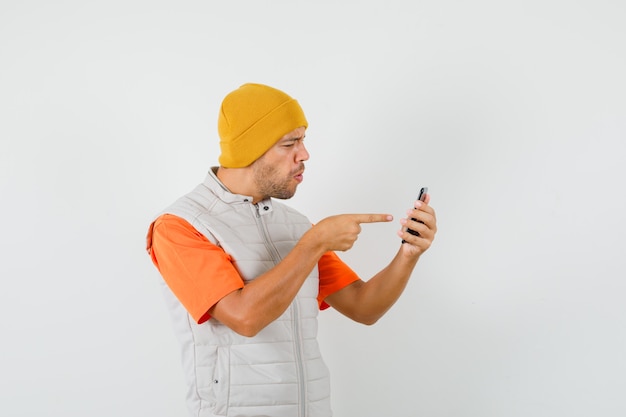 T- 셔츠, 재킷, 모자 전면보기에 휴대 전화에서 가리키는 젊은 남자.