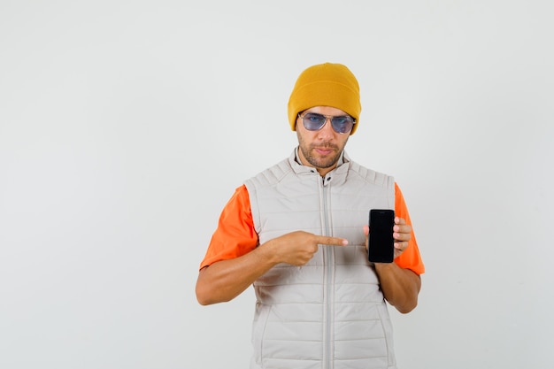 Young man pointing at mobile phone in t-shirt, jacket, hat front view.