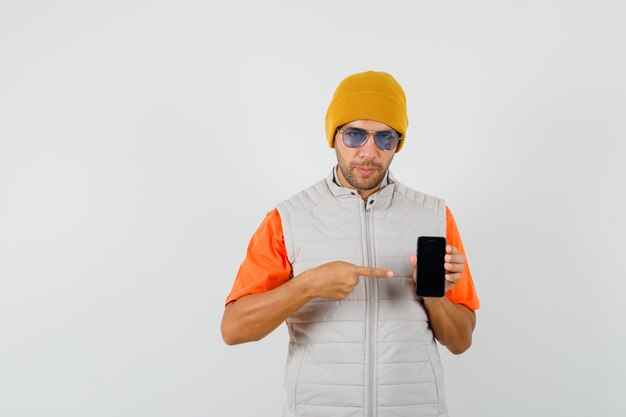 Young man pointing at mobile phone in t-shirt, jacket, hat front view.