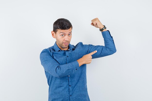 Young man pointing at muscles of arm in blue shirt and looking powerful. front view.