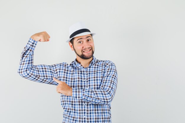 Young man pointing at muscles of arm in checked shirt, hat and looking strong , front view.