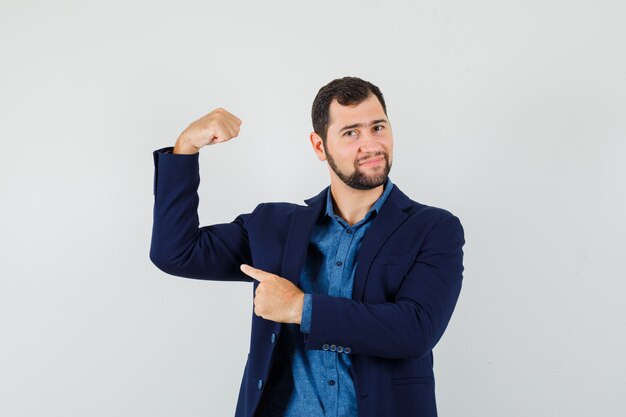 Young man pointing at muscles of arm in shirt, jacket and looking powerful. front view.