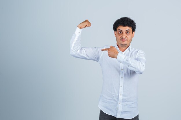 Young man pointing at muscles of arm in white shirt, pants and looking proud. front view.