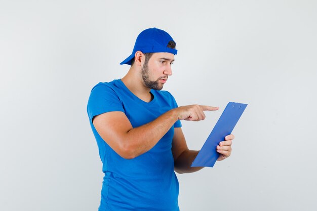 Young man pointing at notes on clipboard in blue t-shirt and cap and looking focused