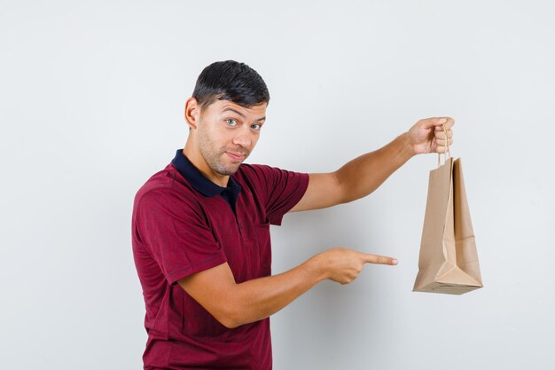 Young man pointing at paper bag in t-shirt and looking curious , front view.