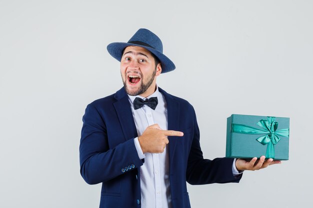 Young man pointing at present box in suit, hat and looking merry , front view.