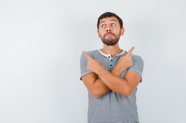 Young man pointing right and left, looking up in striped t-shirt and looking hesitative. front view.