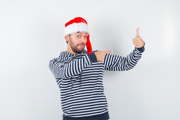 Young man pointing right, showing thumb up in hoodie, Santa hat and looking pleased ,