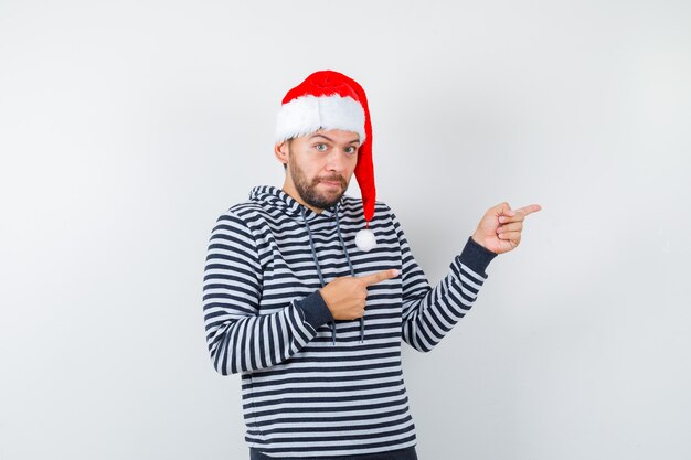 Young man pointing to the right side in hoodie, Santa hat and looking confident ,