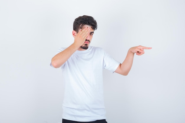 Young man pointing to the right side while covering eye in t-shirt and looking confident