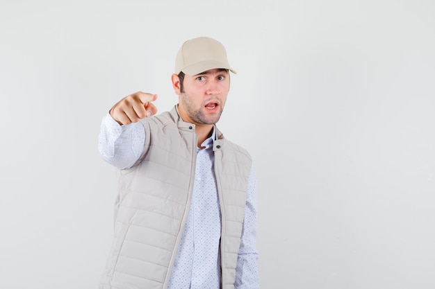 Young man pointing in shirt,sleeveless jacket,cap and looking serious. front view.