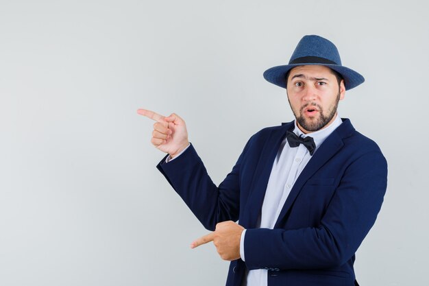 Young man pointing to the side and down in suit, hat and looking confused , front view.