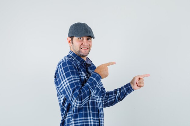 Young man pointing to the side in shirt, cap and looking cheerful. .