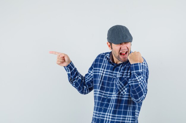 Young man pointing to the side in shirt, cap and looking lucky , front view.