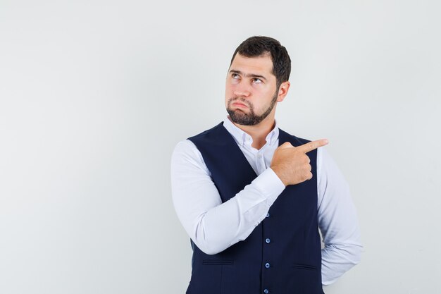 Young man pointing to side in shirt, vest and looking pensive