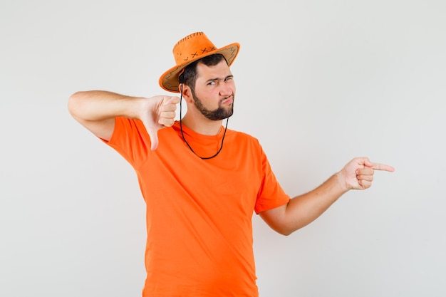 Молодой человек, указывая в сторону, показывая большой палец вниз в оранжевой футболке, шляпе и недовольно выглядел. передний план.