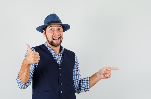 Young man pointing to the side, showing thumb up in shirt, vest, hat and looking glad