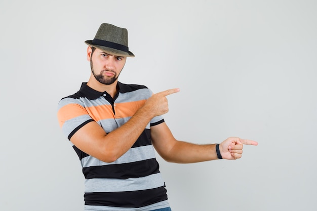 Young man pointing to the side in t-shirt, hat and looking downcast.