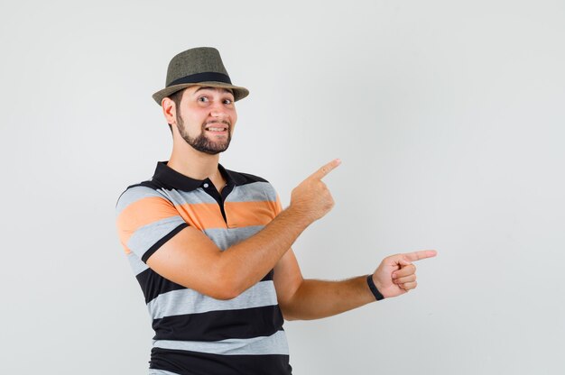 Young man pointing to the side in t-shirt, hat and looking optimistic.