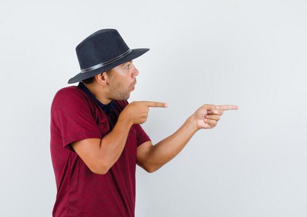 Young man pointing to side in t-shirt, hat and looking wondered. front view.