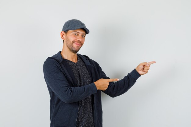 Young man pointing to the side in t-shirt, jacket, cap and looking optimistic.