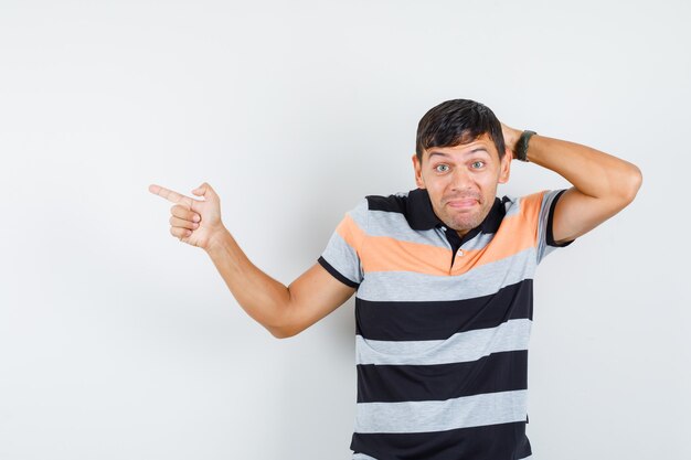 Young man pointing to side with hand behind head in t-shirt and looking curious