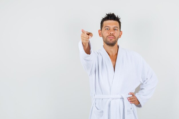 Young man pointing at something away in white bathrobe and looking serious. front view.