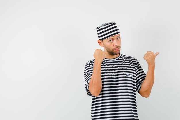 Young man pointing thumb away showing fist in striped t-shirt hat and looking serious 