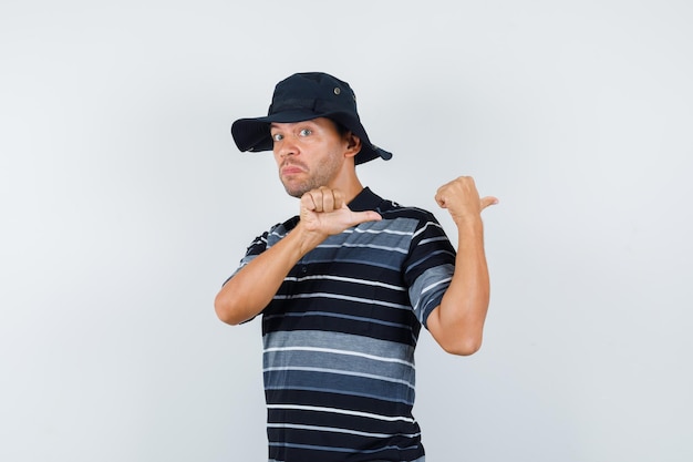 Young man pointing thumbs back in t-shirt, hat and looking irresolute , front view.