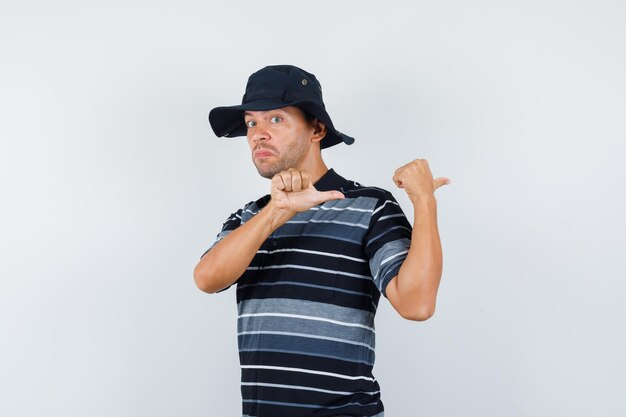 Young man pointing thumbs back in t-shirt, hat and looking irresolute , front view.