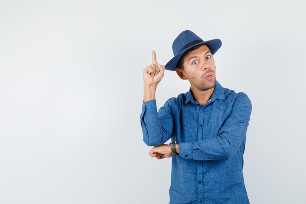 Young man pointing up in blue shirt, hat and looking curious , front view.