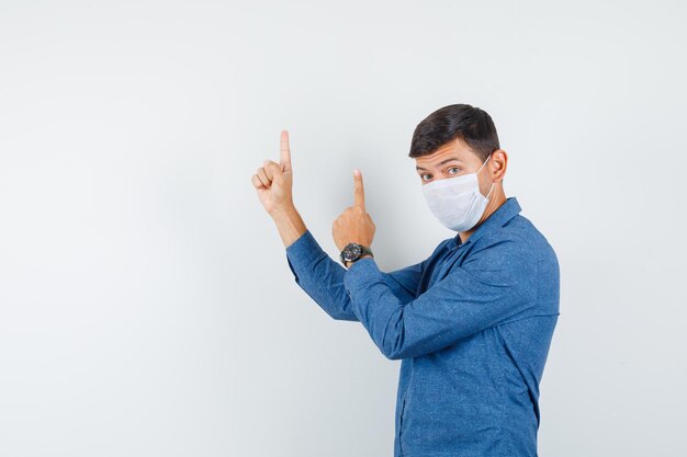 Young man pointing up in blue shirt, mask and looking serious. .