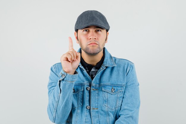 Young man pointing up in cap, t-shirt, jacket and looking curious , front view.