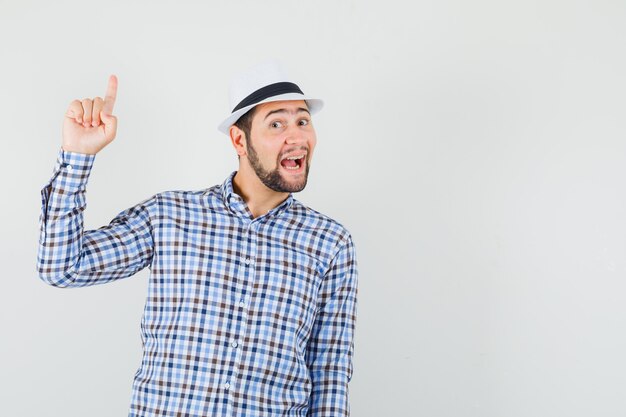 Young man pointing up in checked shirt, hat and looking cheerful.