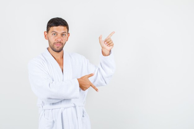 Young man pointing up and down with gun gesture in white bathrobe front view.