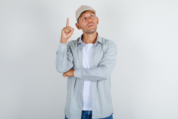 Young man pointing up finger in t-shirt with cap, jeans and looking thoughtful