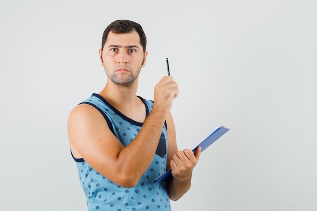 Young man pointing up, holding clipboard in blue singlet and looking serious , front view.