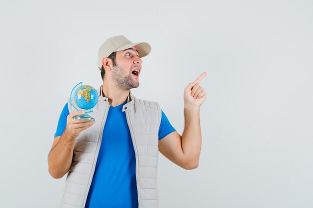 Young man pointing up, holding school globe in t-shirt, jacket, cap and looking happy , front view.