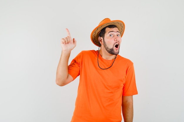 Молодой человек указывая вверх в оранжевой футболке, шляпе и любопытно глядя, вид спереди.
