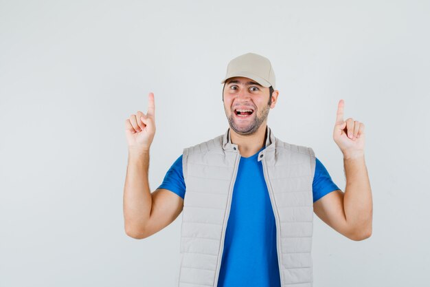 Young man pointing up in t-shirt, jacket, cap and looking merry , front view.