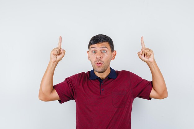 Young man pointing up in t-shirt and looking curious , front view.