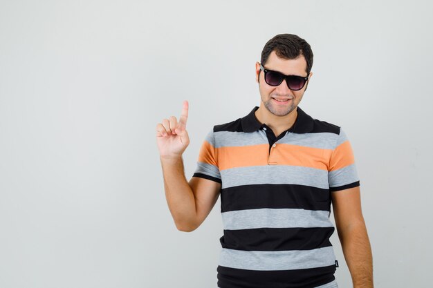 Young man pointing up in t-shirt,sunglasses and looking confident. front view.