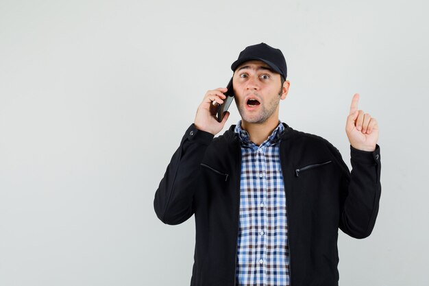 Young man pointing up, talking on mobile phone in shirt, jacket, cap and looking surprised. front view.