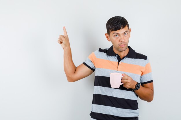 Young man pointing up while holding cup of drink in t-shirt and looking serious